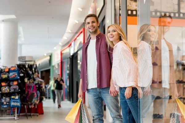 Молодая счастливая пара с бумажными пакетами выходит из магазина в торговом центре — стоковое фото