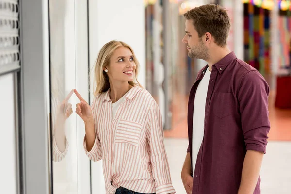 Young woman pointing by finger at shopwindow to boyfriend with shopping bags at mall — Stock Photo