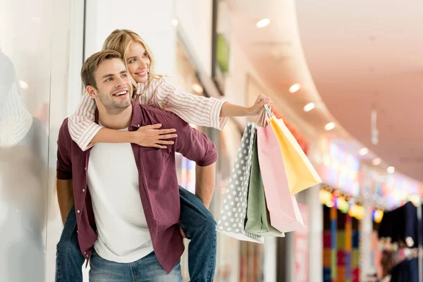 Feliz joven dando paseo a cuestas a su novia con bolsas de compras en el centro comercial - foto de stock