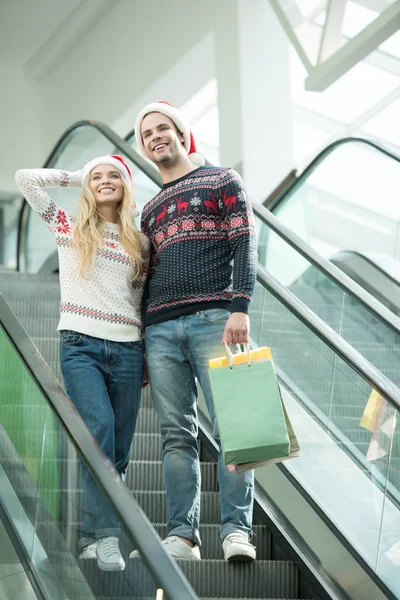 Baixo ângulo de visão de casal de compradores em chapéus de Natal segurando sacos de papéis na escada rolante — Fotografia de Stock