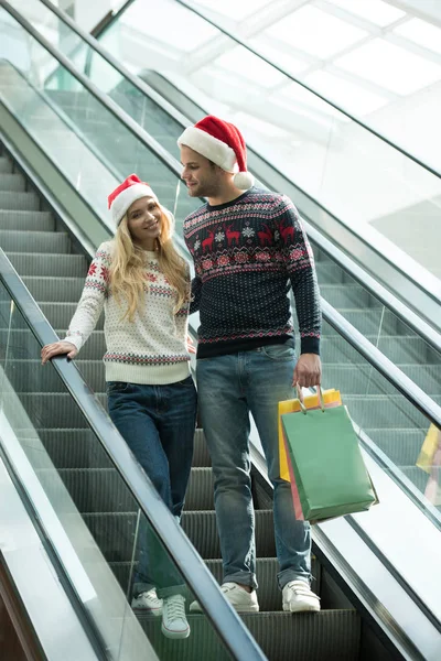 Счастливая молодая пара покупателей в рождественских шляпах с бумажными пакетами на эскалаторе — стоковое фото