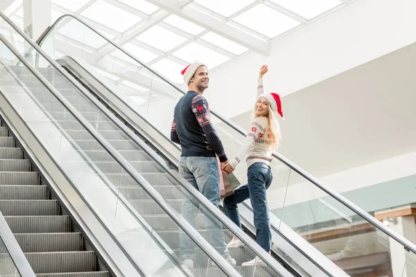 Junge Frau mit Weihnachtsmütze mit erhobenen Armen blickt in die Kamera, während ihr Freund in der Nähe auf der Rolltreppe steht — Stockfoto