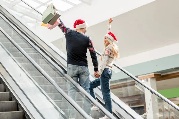 Rückansicht eines Shopperpaares in Weihnachtsmützen mit erhobenen Armen, die Papiertüten auf der Rolltreppe halten — Stockfoto