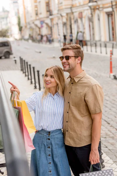 Mujer joven sonriente con bolsas de papel señalando con el dedo al novio en gafas de sol en los escaparates de la calle de la ciudad - foto de stock
