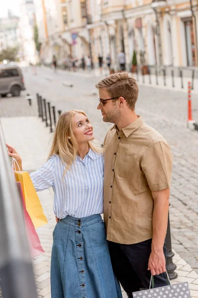 Happy woman with paper bags pointing by finger to boyfriend in sunglasses at shopwindows on city street — Stock Photo