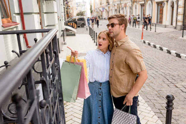 Mujer conmocionada con bolsas de papel apuntando con el dedo al novio en gafas de sol en los escaparates de la calle de la ciudad - foto de stock