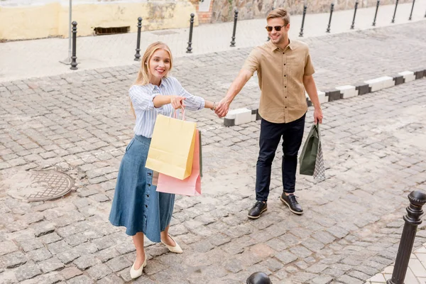 Visão de alto ângulo da jovem com sacos de papel apontando por dedo para namorado em óculos de sol em shopwindows na rua da cidade — Fotografia de Stock
