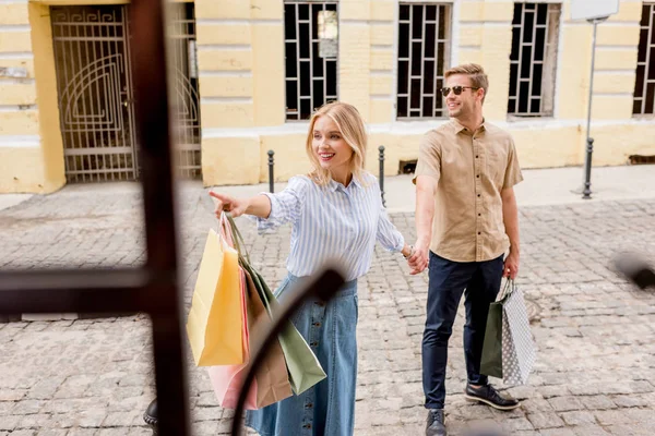 Усміхнена молода жінка з паперовими мішками вказує пальцем на хлопця в сонцезахисних окулярах на вітринах магазинів на вулиці міста — стокове фото