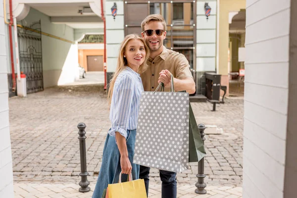 Stylish young man in sunglasses holding shopping bags and pointing by finger to girlfriend on street — Stock Photo