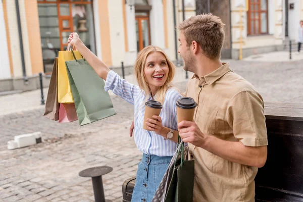 Young woman with coffee and shopping bags pointing by hand to boyfriend standing near at street — Stock Photo