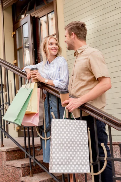 Smiling young couple with shopping bags and paper cups of coffee standing on staircase at urban street — Stock Photo