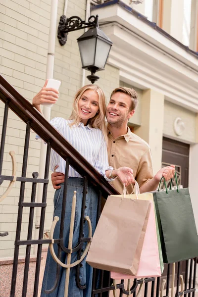 Couple élégant avec sacs à provisions prendre selfie sur smartphone près du magasin à la rue — Photo de stock