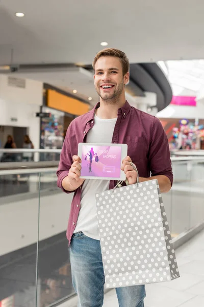 Молодой красивый мужчина с бумажным пакетом, показывающий цифровой планшет с интернет-магазинами на экране в торговом центре — стоковое фото