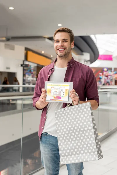 Smiling young man with paper bag showing digital tablet with aliexpress website on screen at shopping mall — Stock Photo