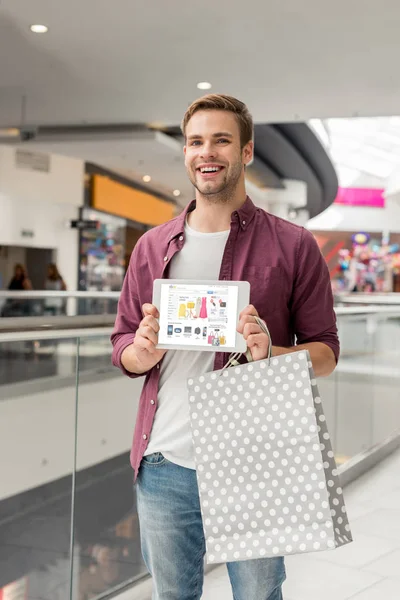 Hombre sonriente guapo con bolsa de papel que muestra la tableta digital con el sitio web de eBay en la pantalla en el centro comercial - foto de stock