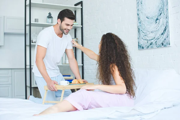 Smiling young man bringing breakfast to beautiful girlfriend in bed — Stock Photo