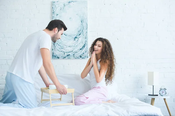 Smiling young man bringing breakfast to beautiful surprised girlfriend in bed — Stock Photo