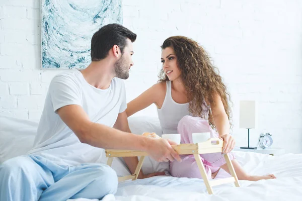 Красивая молодая пара в пижаме улыбается друг другу во время завтрака вместе в постели — стоковое фото