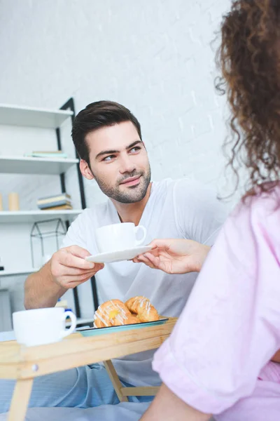 Handsome young man holding cup of coffee and looking at girlfriend in  bedroom — Stock Photo