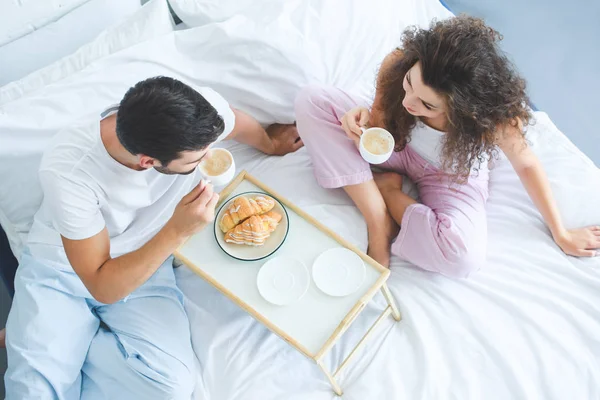 Vista aérea de pareja joven en pijama tomando café y desayunando en la cama - foto de stock