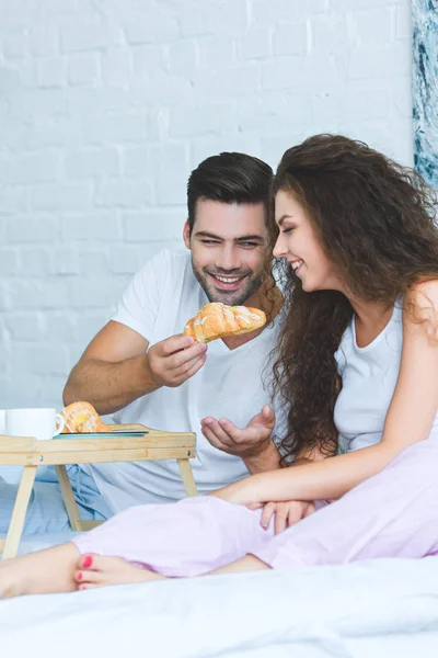 Handsome smiling young man feeding beautiful girlfriend with croissant in bedroom — Stock Photo