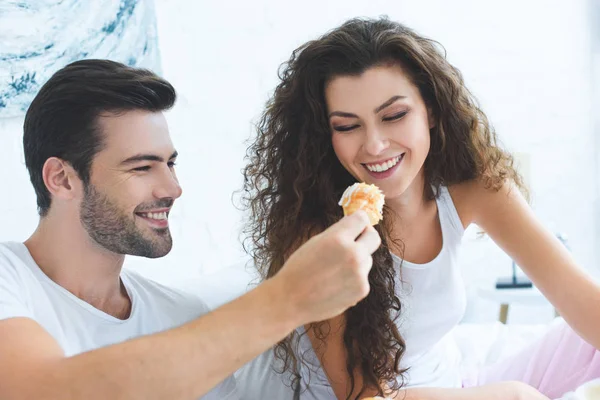 Smiling young man feeding happy girlfriend with croissant for breakfast in bedroom — Stock Photo