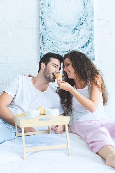 Красивая счастливая молодая женщина кормит улыбающегося парня круассаном в постели — стоковое фото