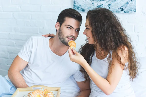 Belle jeune femme nourrissant petit ami souriant avec croissant au lit — Photo de stock