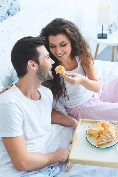 Счастливая молодая женщина кормит улыбающегося парня круассаном в постели — стоковое фото