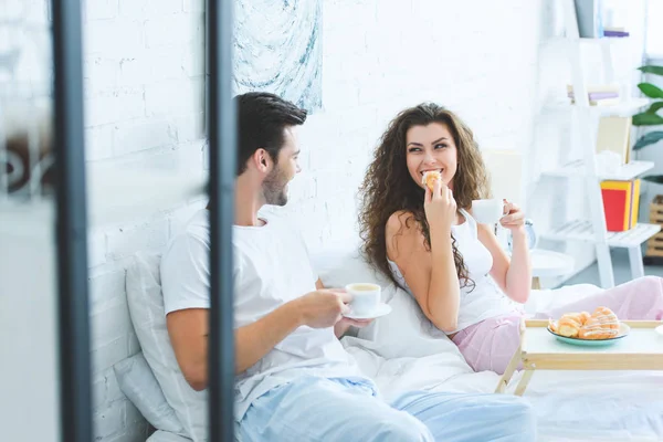 Selective focus of happy young couple in pajamas having breakfast and smiling each other in bedroom — Stock Photo