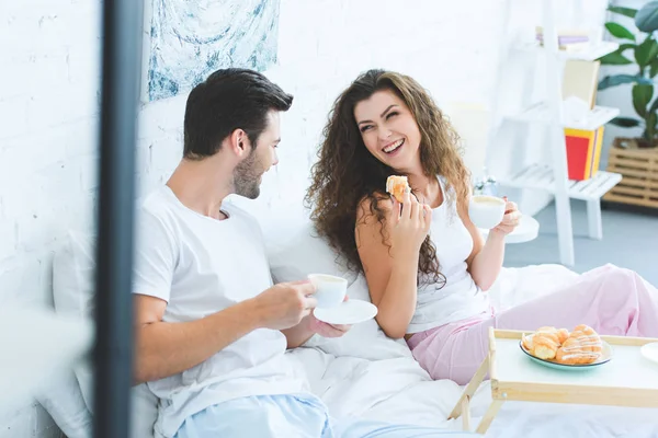 Счастливая молодая пара в пижаме завтракает и улыбается друг другу в спальне — стоковое фото