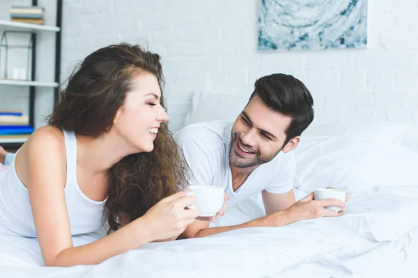 Cheerful young couple in pajamas holding cups of coffee and lying on bed — Stock Photo
