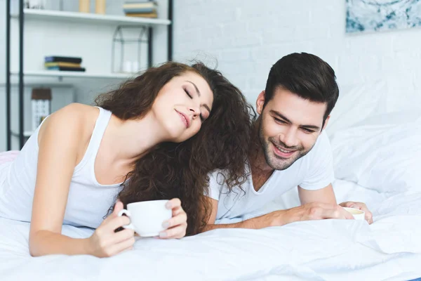 Hermosa sonriente joven pareja sosteniendo tazas de café y acostado en la cama - foto de stock