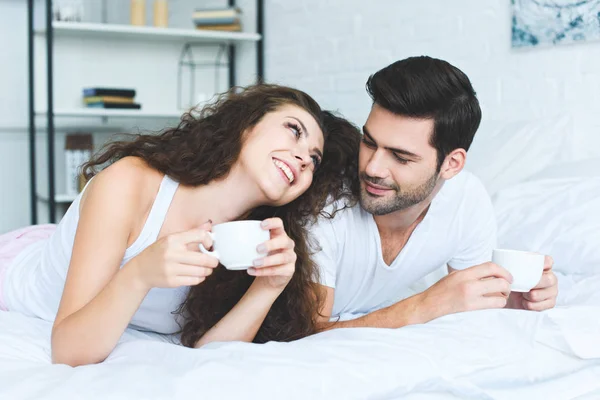 Beautiful happy young couple holding cups of coffee and lying on bed — Stock Photo