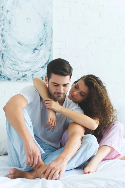 Beautiful young woman hugging handsome husband while sitting together on bed — Stock Photo