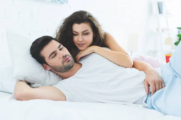 Beautiful smiling young woman looking at sleeping boyfriend in bedroom — Stock Photo