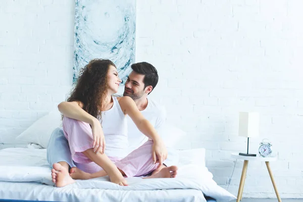 Feliz joven pareja en pijama sentado en la cama y sonriéndose mutuamente - foto de stock