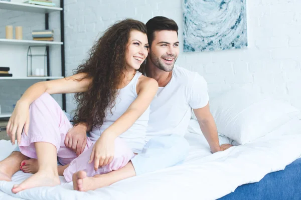 Glückliches junges Paar im Pyjama, das zusammen auf dem Bett sitzt und wegschaut — Stockfoto