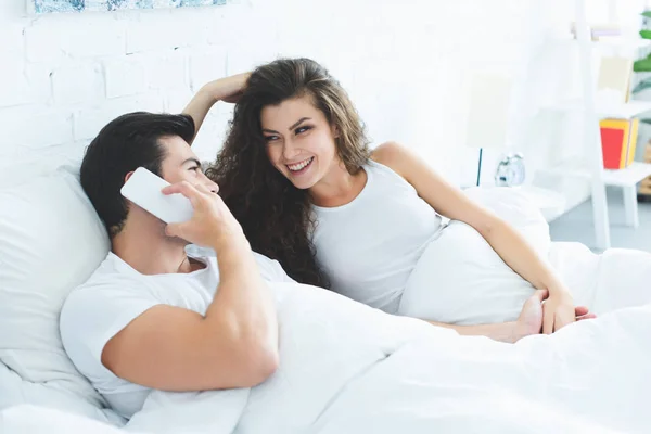 Smiling young woman looking at boyfriend talking by smartphone in bed — Stock Photo