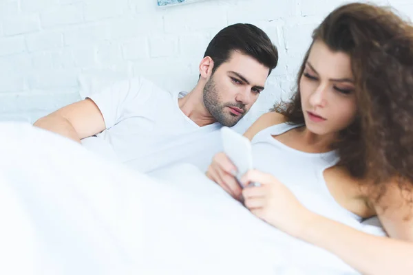 Jeune un regardant petite amie en utilisant smartphone au lit, concept de difficultés relationnelles — Photo de stock