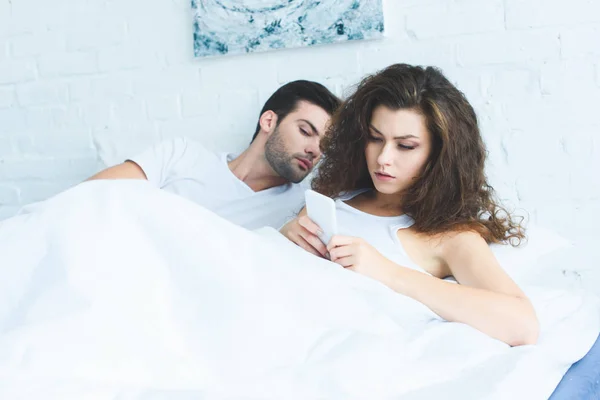Серьезная молодая женщина с помощью смартфона во время лежания с парнем в постели — стоковое фото