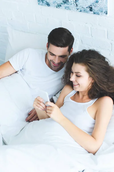 Vista de ángulo alto de hermosa pareja joven y feliz utilizando el teléfono inteligente juntos en la cama - foto de stock
