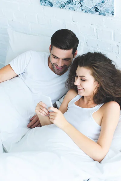 Visão de alto ângulo de belo casal jovem feliz usando o smartphone juntos na cama — Fotografia de Stock