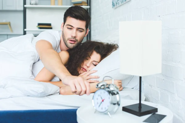 Sleepy young man reaching for alarm clock while sleeping with girlfriend in bed — Stock Photo