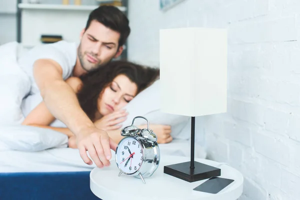 Young man reaching for alarm clock while sleeping with girlfriend in bed — Stock Photo