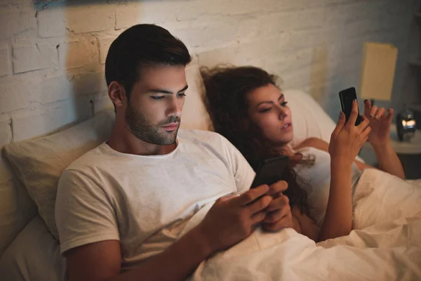 Junges Paar nutzt Smartphones, während es nachts im Bett liegt — Stockfoto