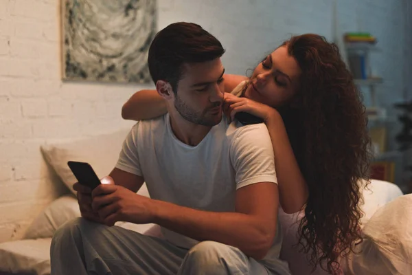 Attractive young woman looking at boyfriend using smartphone on bed at night — Stock Photo