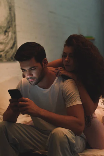 Young woman looking at boyfriend using smartphone on bed at night — Stock Photo