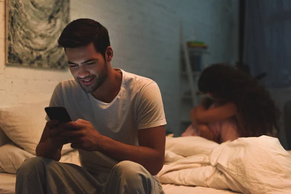 Homme utilisant smartphone tandis que la petite amie bouleversée assis sur le lit la nuit, concept de difficultés relationnelles — Photo de stock
