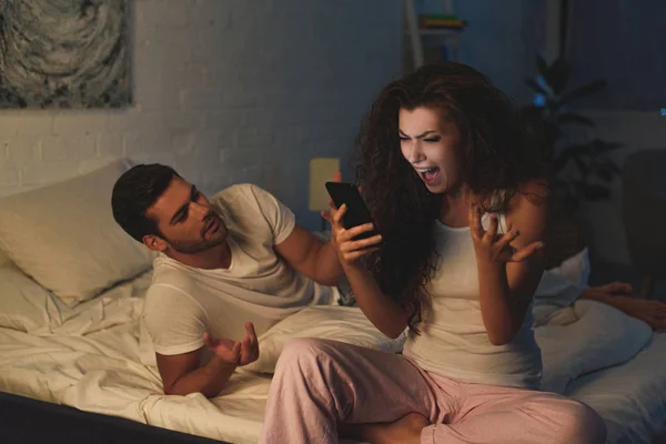 Mujer joven enojada usando un teléfono inteligente y gritando mientras su novio está acostado en la cama por la noche - foto de stock